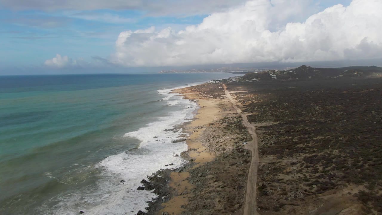 Aerial view of Punta Gorda coastline with blue-green ocean on the left, white surf reaching a sandy and rocky shore, a dirt road inland, distant hills, large clouds, and no visible people.