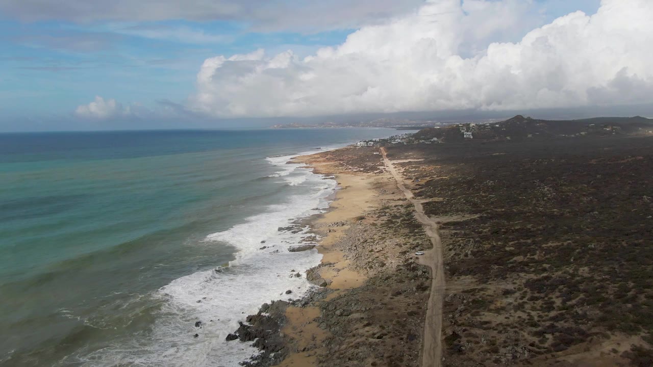 Drone view along a beach where waves break beside a dirt coastal road and scrubland.