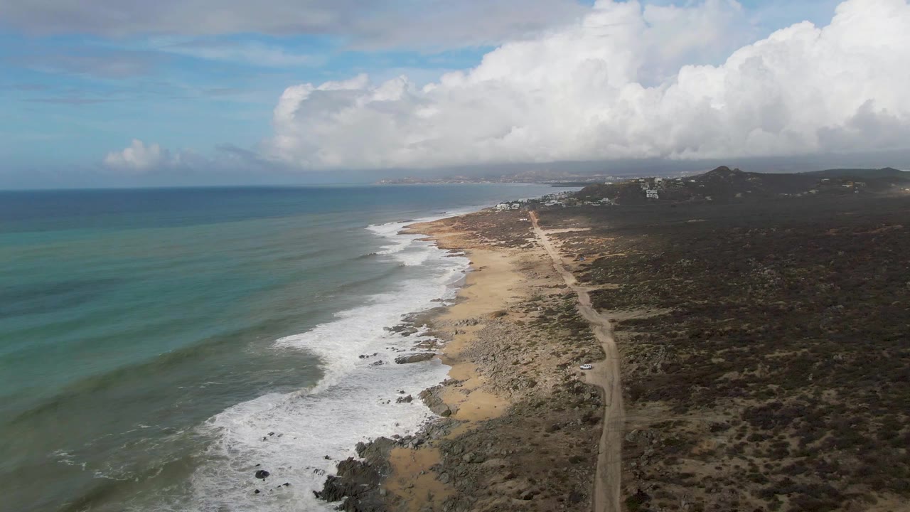 Oblique aerial view of the Punta Gorda shore with long wave bands offshore, white water on the sand, a tan road cutting through dark scrubland, hillside terrain beyond, and no visible people.