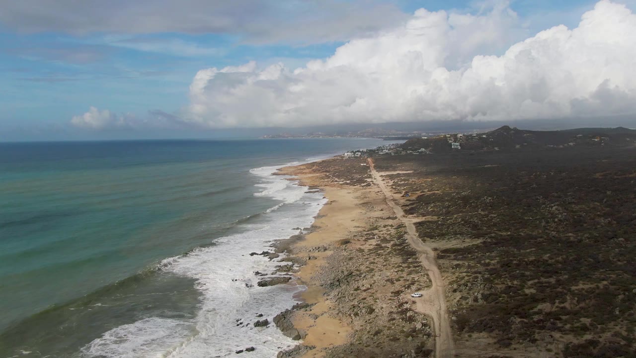 Aerial coastline with ocean, surf, beach, dirt road, scrubland, and clouds.