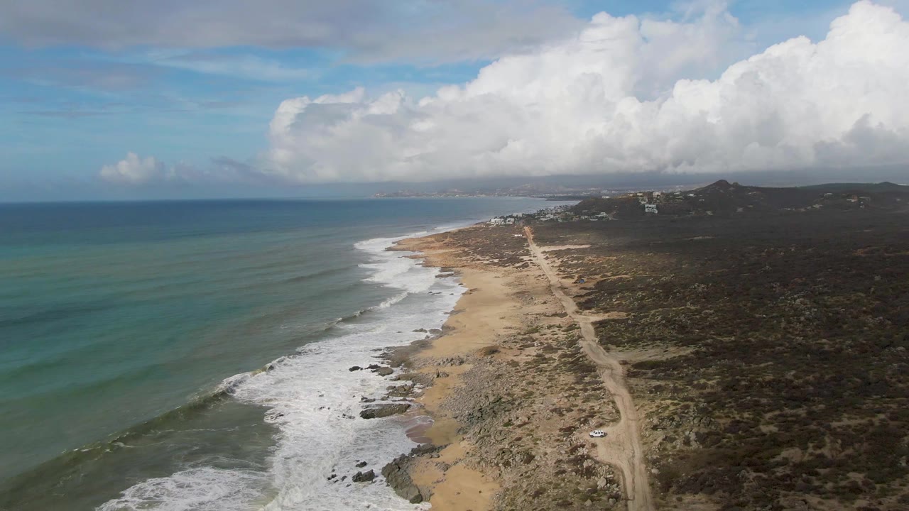 Drone view over Punta Gorda, Baja California Sur showing the shoreline angled into the distance, white surf along the beach, a pale dirt road near the water, scrub-covered land, cloud shadow, and no visible people.