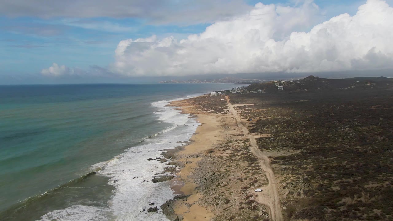 High aerial frame of Punta Gorda, Baja California Sur with turquoise water shifting to deeper blue offshore, surf lines on the beach, the dirt road tracing the coast, darker scrubland, low hills, bright clouds, and no visible people.