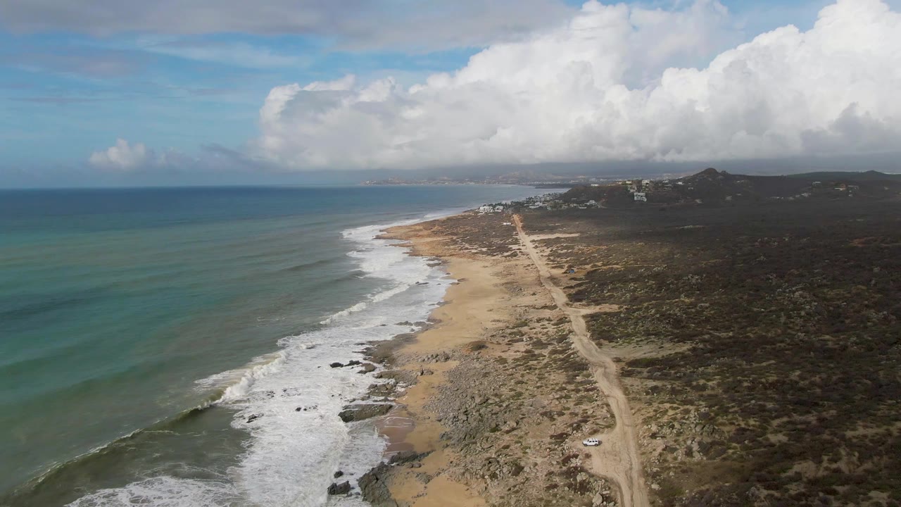 Aerial frame over Punta Gorda, Baja California Sur with foam spreading along the lower shoreline, the dirt road visible beside scrubland, small vehicles on the track, hazy distant coast, broad cloud cover, and no visible people.