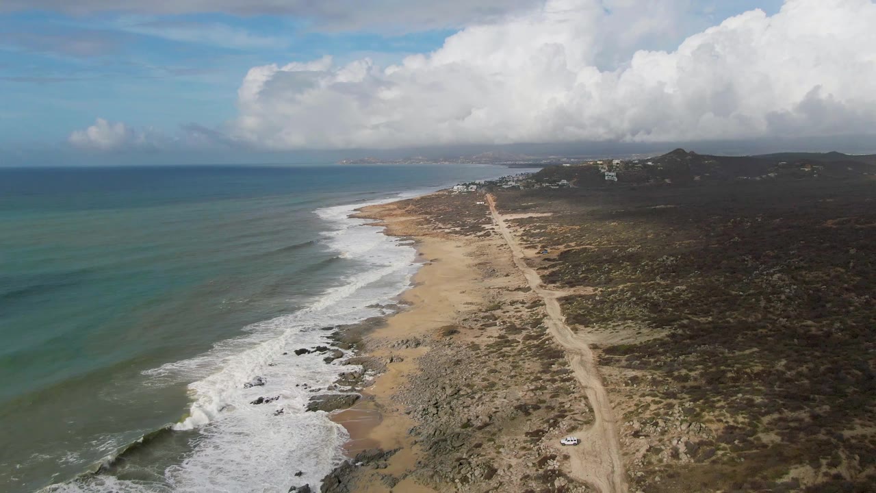 Aerial frame over Punta Gorda, East Cape, Baja California Sur, with green-blue ocean on the left, white surf along a sandy rocky beach, a dirt road through scrubland, and no visible people.