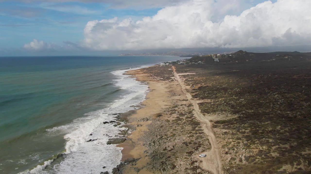 Aerial view in Punta Gorda, East Cape, Baja California Sur, showing the shoreline curving away, waves breaking in bands, the dirt road near the beach, scattered distant structures, and no visible people.