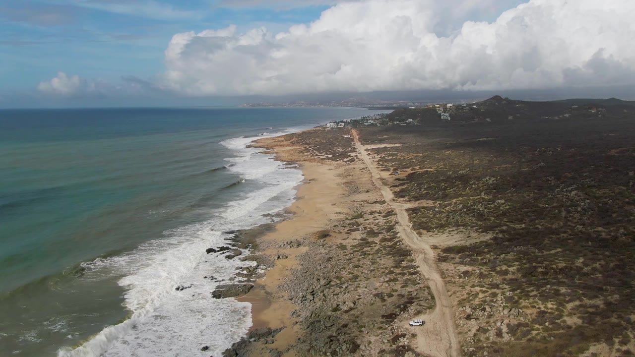 Wide aerial frame over Punta Gorda, East Cape, Baja California Sur, with a long dirt track beside the beach, white foam along the waterline, dark scrubland to the right, distant hills, and no visible people.
