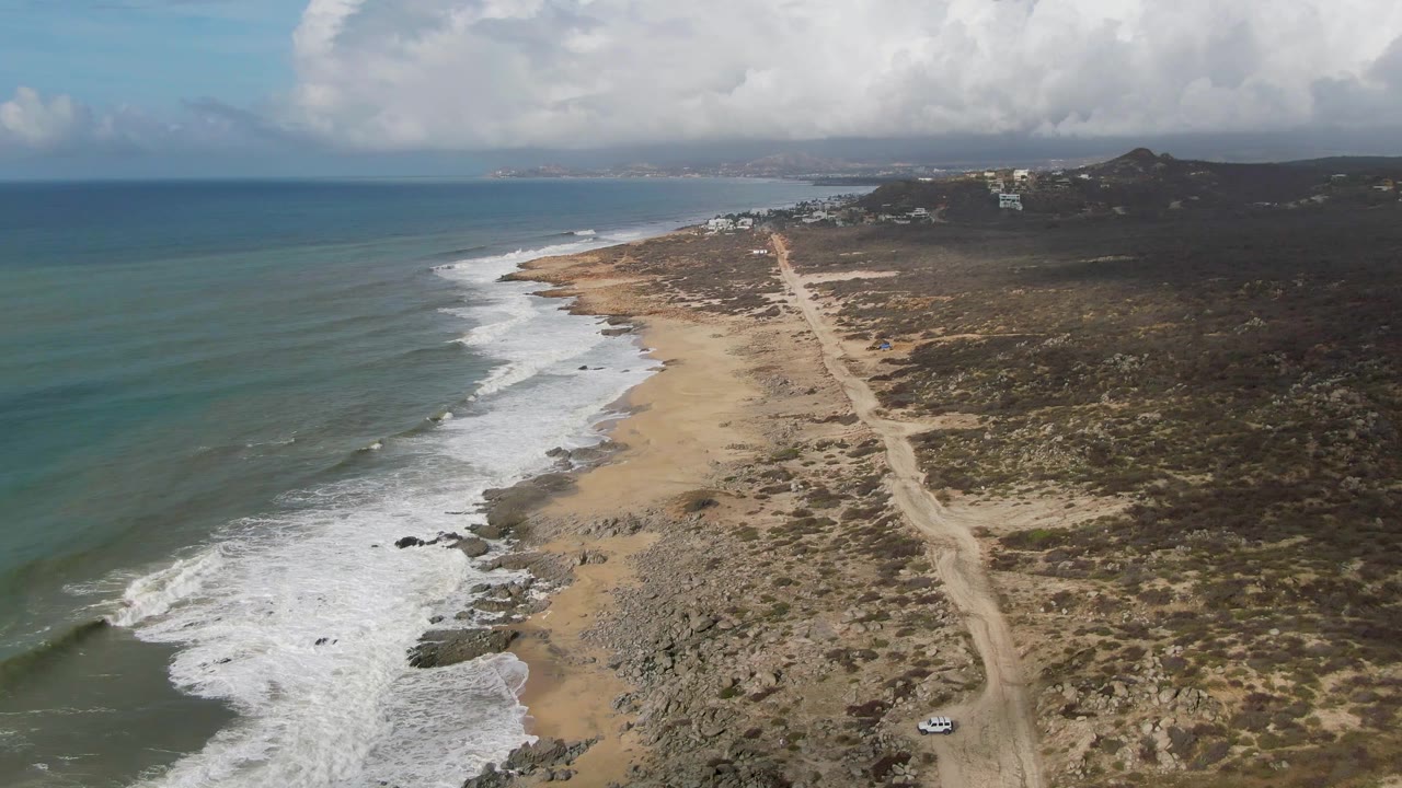 Aerial frame over Punta Gorda, Baja California Sur, with blue-green ocean, white surf, sandy rocky shore, a dirt coastal road, a small white car near the lower road area, and no visible people.