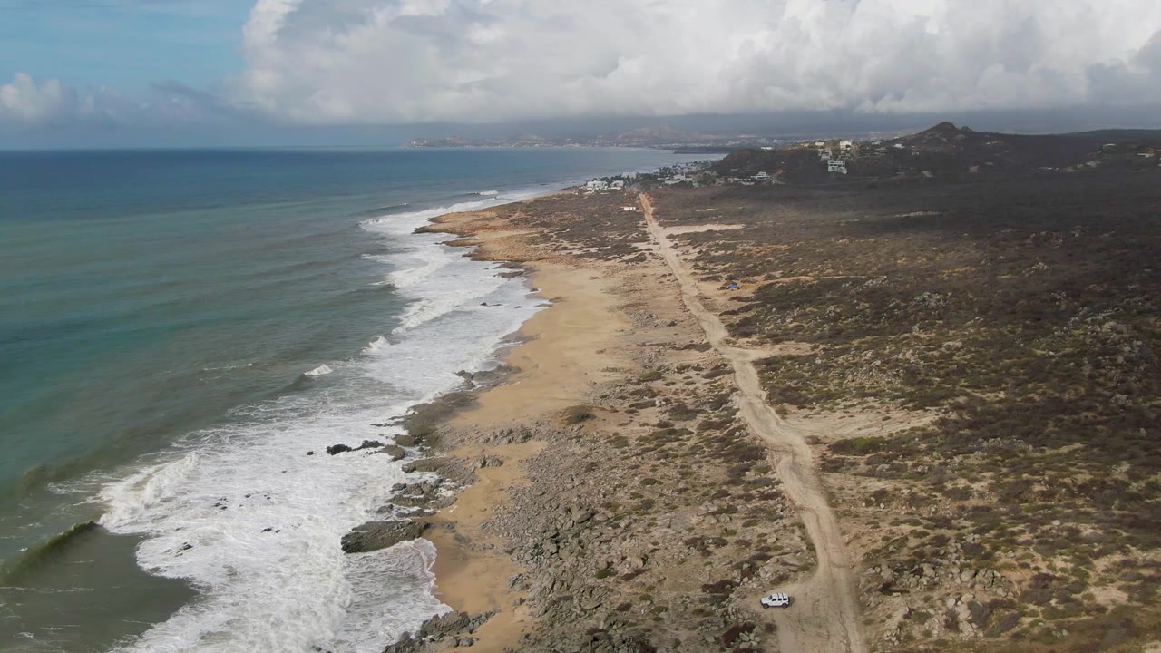 Aerial view of Punta Gorda coastline with ocean on the left, broken white waves along the sandy and rocky shore, a dirt road through scrubland, distant hills, and no visible people.