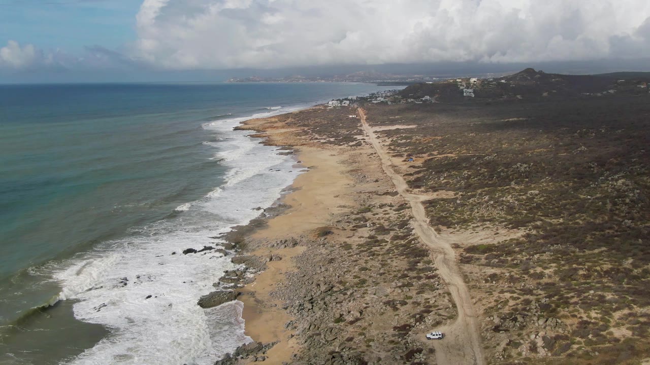 Drone-style aerial frame over Punta Gorda with the coastline angling into the distance, surf bands on the left, a pale dirt road near center, dark scrubland to the right, and no visible people.