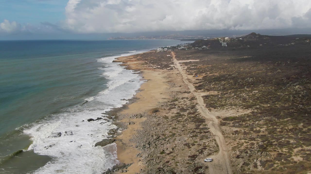 Coastal aerial with surf at left and a dirt road crossing dry scrubland.