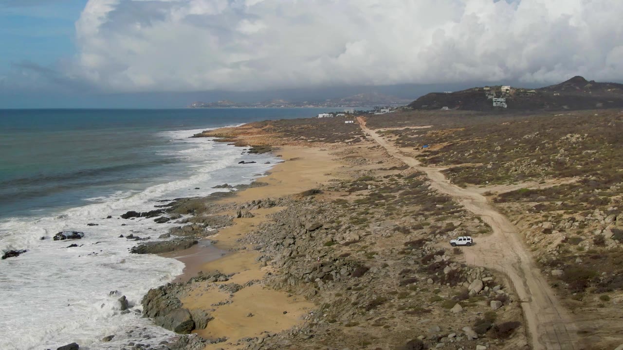 At Punta Gorda in Baja California Sur, a white car appears on the dirt road above a rocky sandy beach, with ocean waves at left, scrub hills at right, cloudy sky, and no visible people.