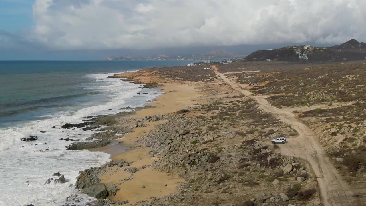 Aerial view of a white car on dirt road near a rocky beach.