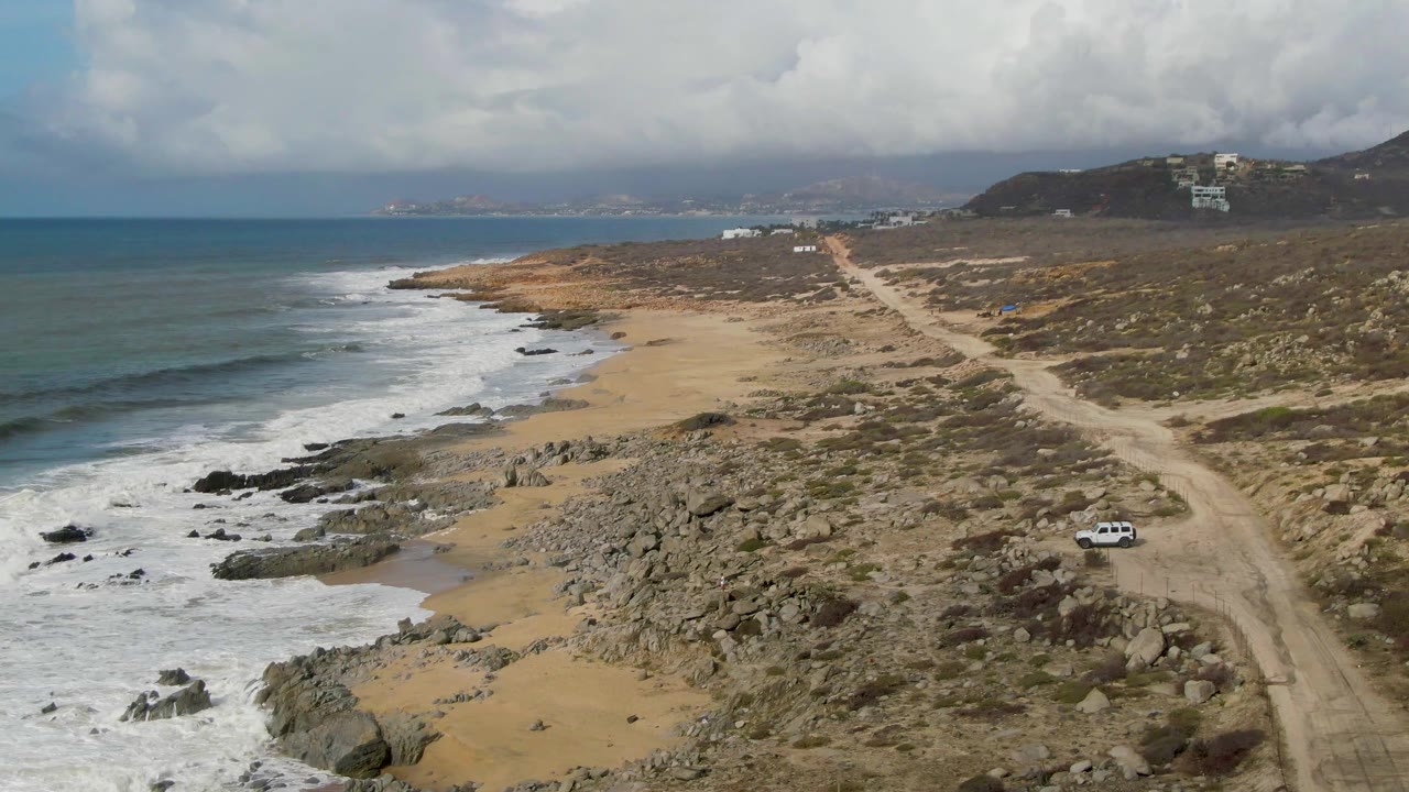 At Punta Gorda in Baja California Sur, the white car sits lower in the frame on the dirt road, with layered surf and scattered black rocks along the beach and no visible people.