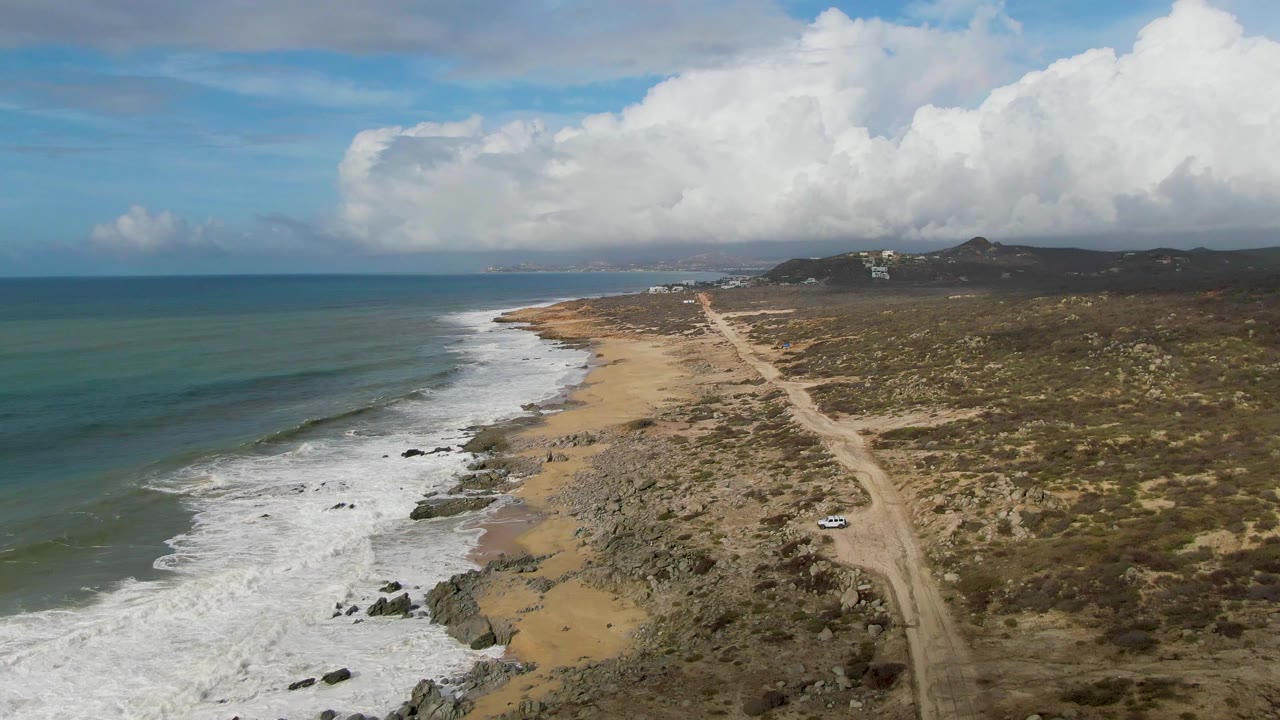 Aerial frame over Punta Gorda, East Cape, Baja California Sur, with surf breaking along the rocky beach and a small white car on the dirt road, no visible people.