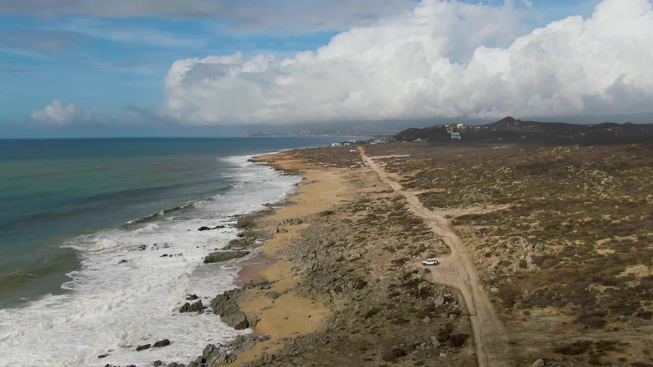 Coastal drone view with ocean surf, rocky beach, dirt road, and a white car.