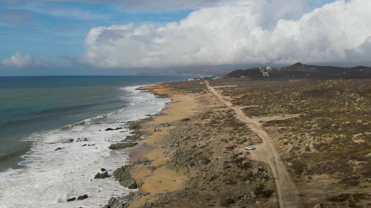 Aerial view in Punta Gorda, Baja California Sur, looking along the shoreline where the road runs beside rocky sand, a white car remains near the lower right, and no people are visible.
