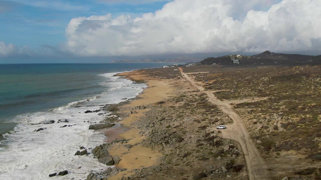 Aerial coastline with waves, rocks, sandy beach, dirt road, and small white car.