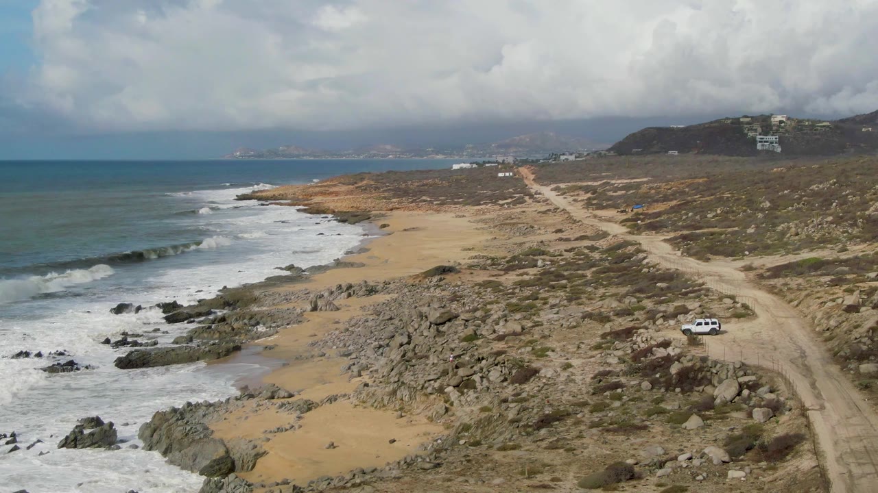 Aerial frame from Punta Gorda, Baja California Sur showing a white car on a dirt road beside rocky sand and breaking ocean waves, with no visible people.
