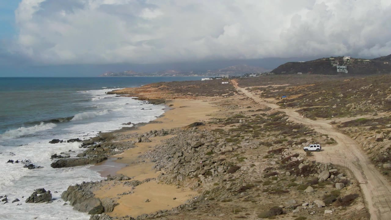 White car on a coastal dirt track above waves and rocks under clouds.