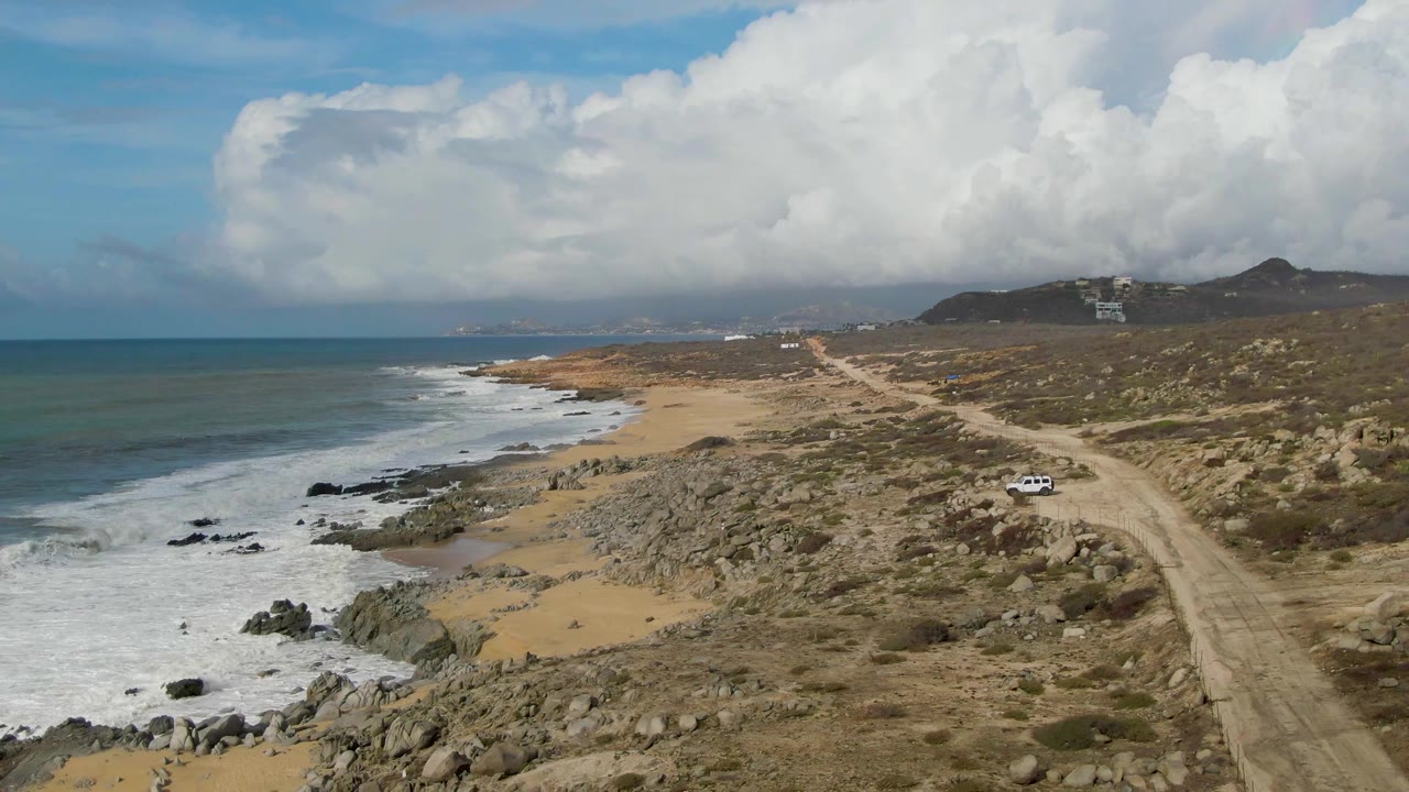 Aerial view in Punta Gordo, Baja California Sur, with ocean waves breaking along a rocky sandy beach, a dirt road inland, low scrub, hills, large clouds, and no visible people.