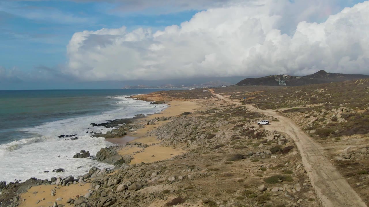 Punta Gordo aerial frame showing the dirt road curving through scrubland, a white vehicle near the road, waves washing over rocks, distant hills, broad clouds, and no visible people.