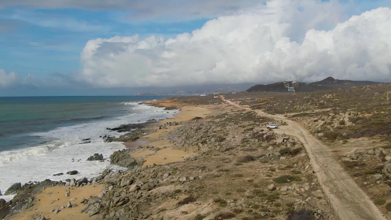 A wide Punta Gordo coastal view with the ocean on the left, white surf crossing dark rocks, a sandy beach strip, a dirt road through dry scrub, vehicles on the track, hills, clouds, and no visible people.