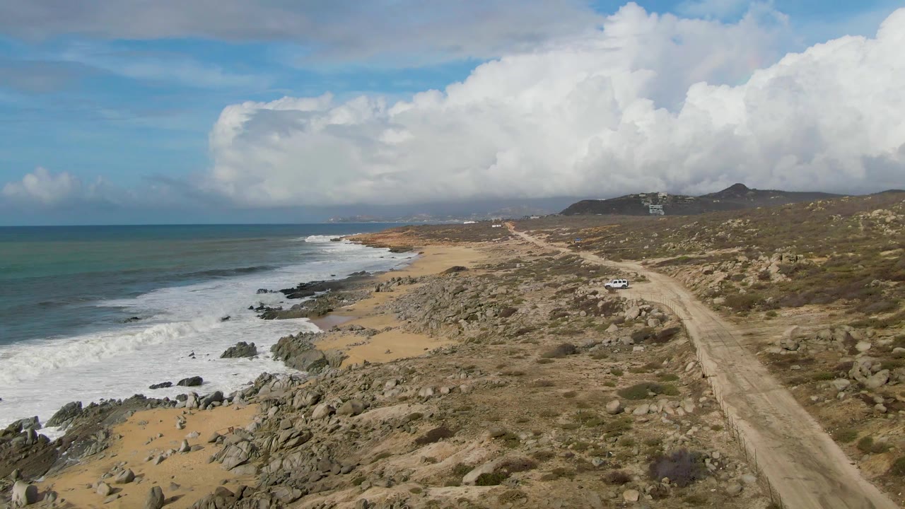 Surf and rocky beach beside a dirt road under a cloud-filled sky.