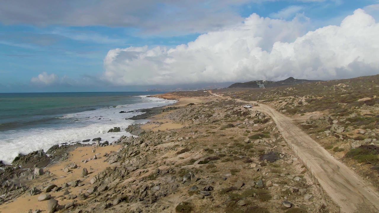 At one second, the Punta Gorda coastal road remains centered while white waves spread across dark shoreline rocks and the cloudy sky brightens the dry scrubland, with no visible people.