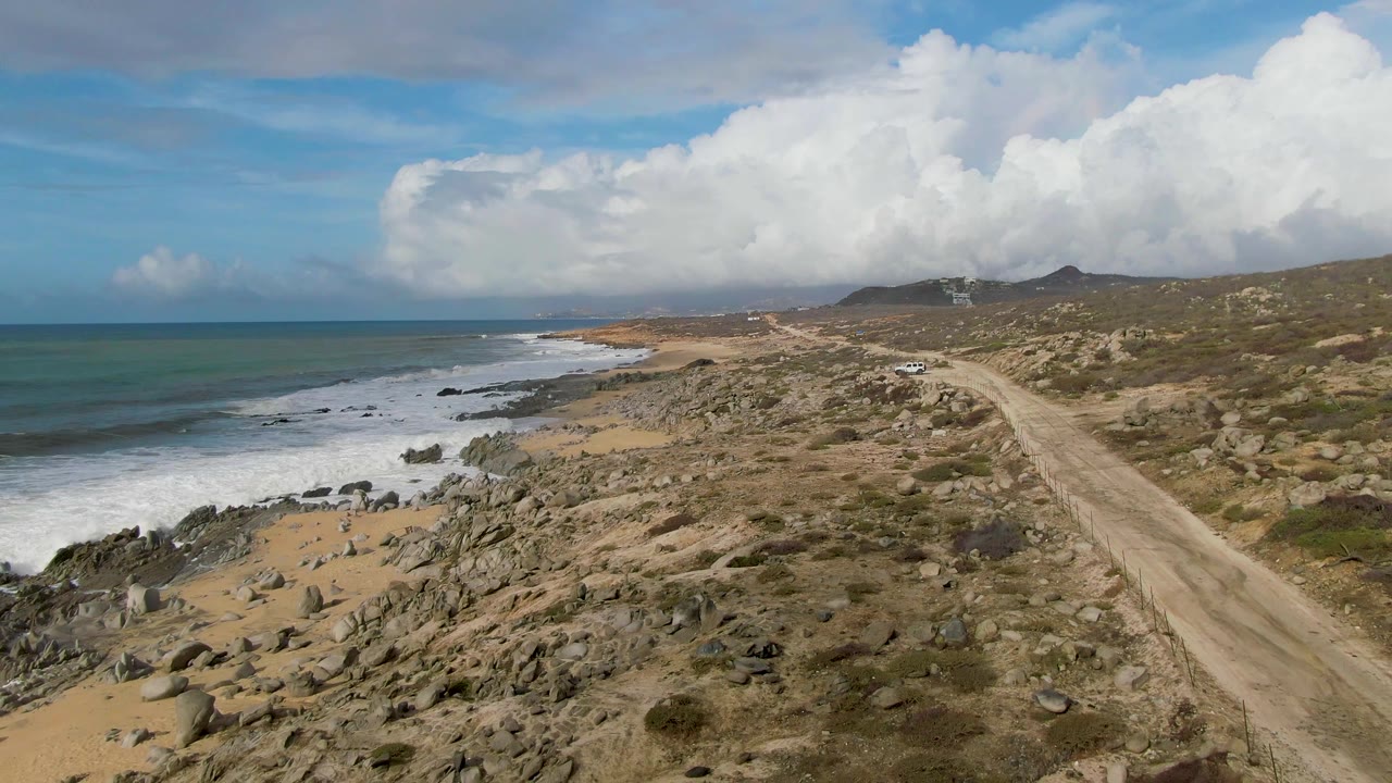 Mid-clip frame in Punta Gorda, Baja California Sur, with the dirt road leading through rocky scrub, surf curling over the beach and rocks, low hills in the distance, and no visible people.