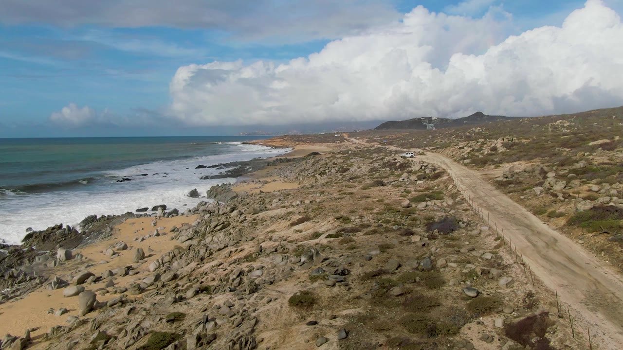 At three seconds, the elevated view looks farther along the Punta Gorda shoreline as the dirt road tracks through dry terrain beside foamy surf and dark rocks, with no visible people.