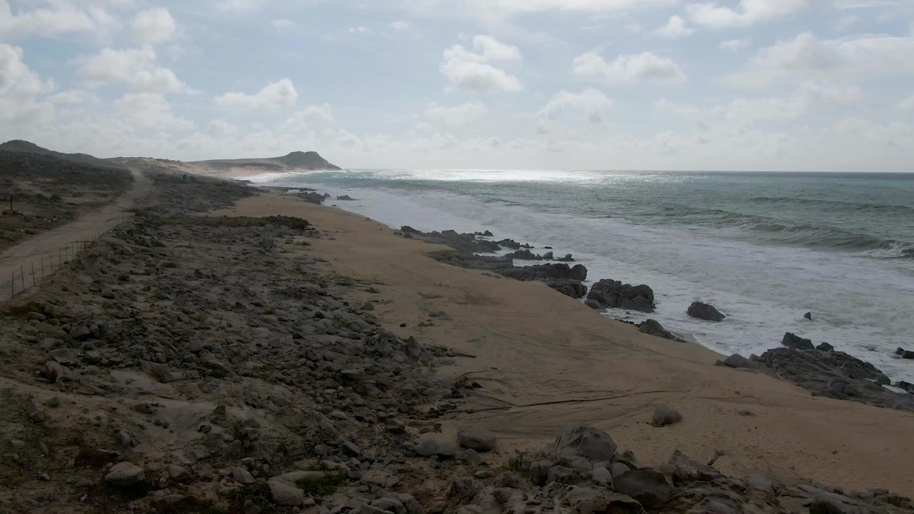 Elevated view of Punta Gorda East Cape shoreline in Baja California Sur, with dark rocks, sand, rolling surf, distant low hills, and no visible people.