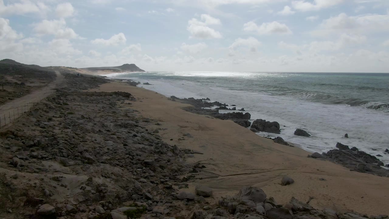 Aerial-looking frame of Punta Gorda East Cape with foamy surf spreading along the rocks, a sandy beach below, low hills in the distance, and no visible people.