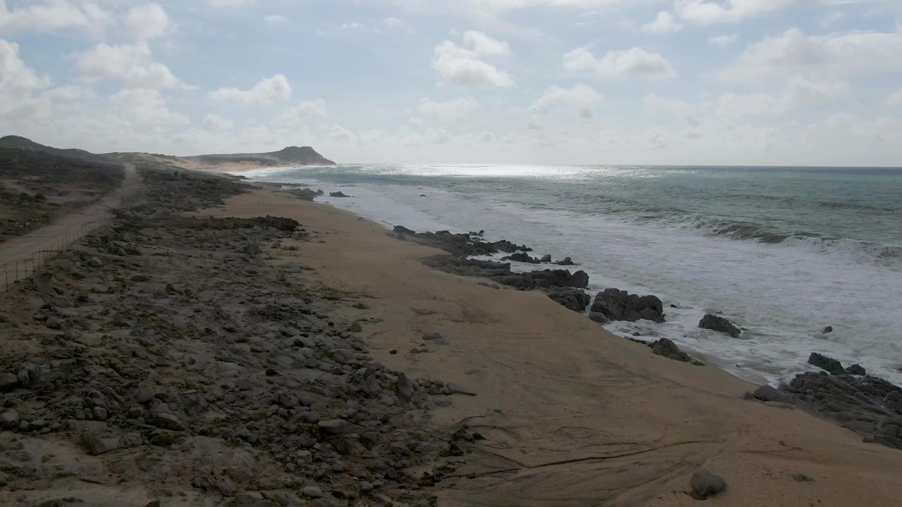 Punta Gorda East Cape shoreline with a broad band of whitewater near the rocks, sandy beach in the lower frame, open ocean to the right, and no visible people.