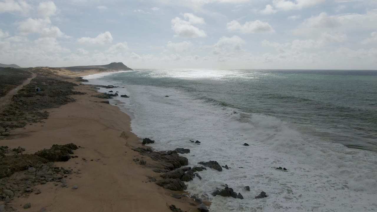 Wide aerial frame over Punta Gorda, Baja California Sur, with the surf band pushing farther along the sandy shore, sun glare on the water, rocks at the waterline, and no visible people.