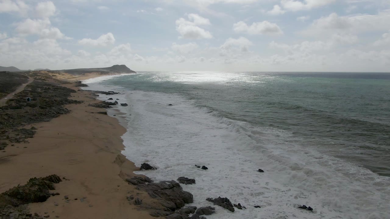 Aerial view of Punta Gorda coastline with white breakers covering more of the beach edge, dark rocks interrupting the foam, blue-green ocean to the right, and no visible people.
