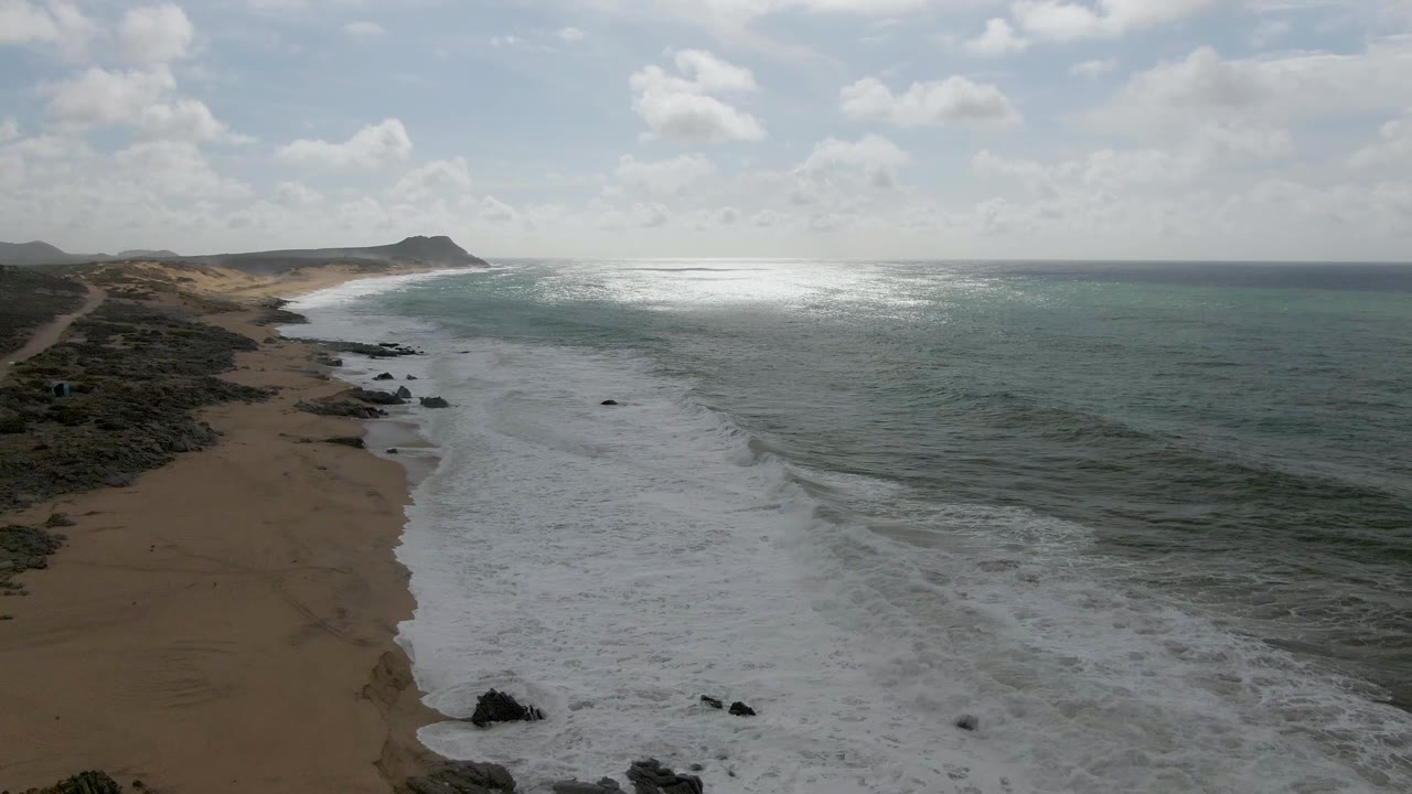 Aerial frame over Punta Gorda, Baja California Sur, with foamy water filling the lower foreground, the sandy beach running up the left side, open ocean stretching right, and no visible people.