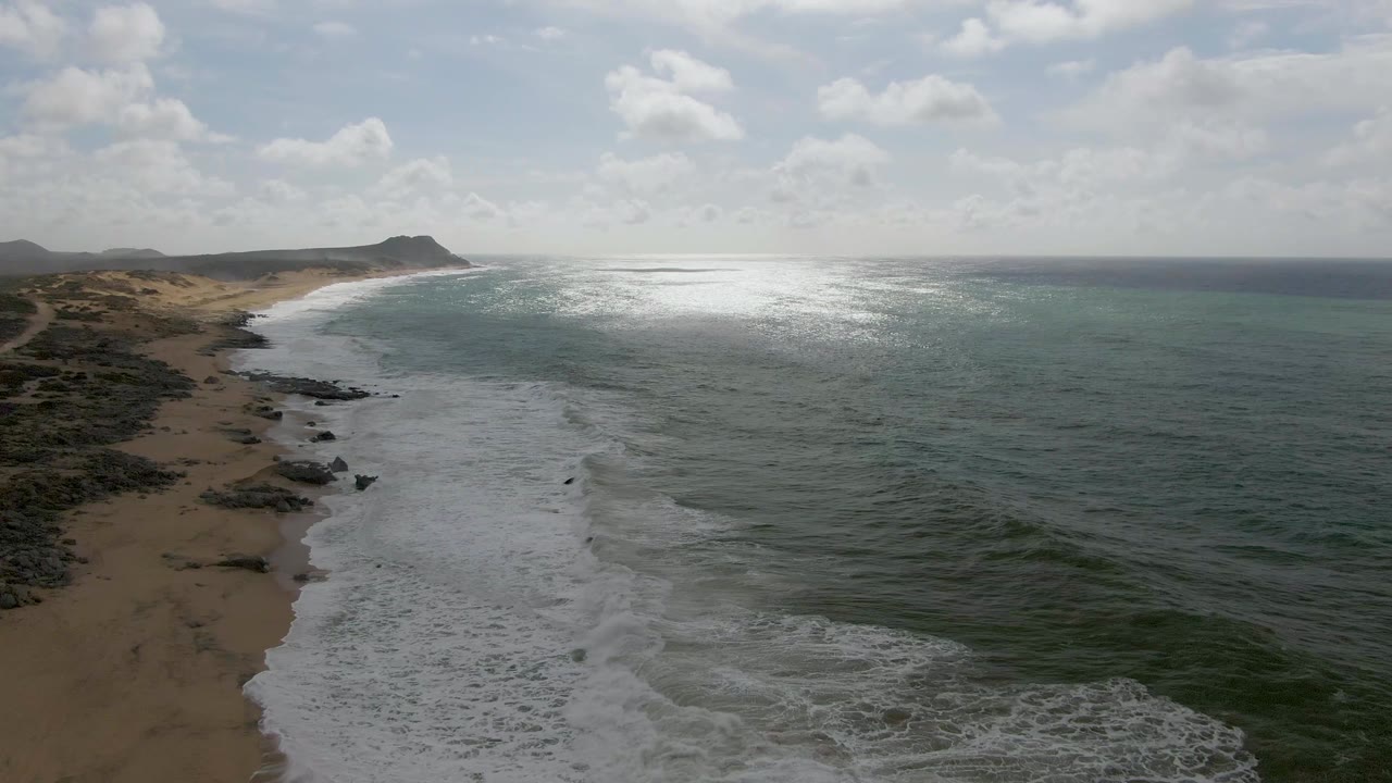 Aerial frame of Punta Gorda East Cape shoreline with white waves reaching the sandy beach, dark rocks near the waterline, green-blue ocean, distant headland, partly cloudy sky, and no visible people.