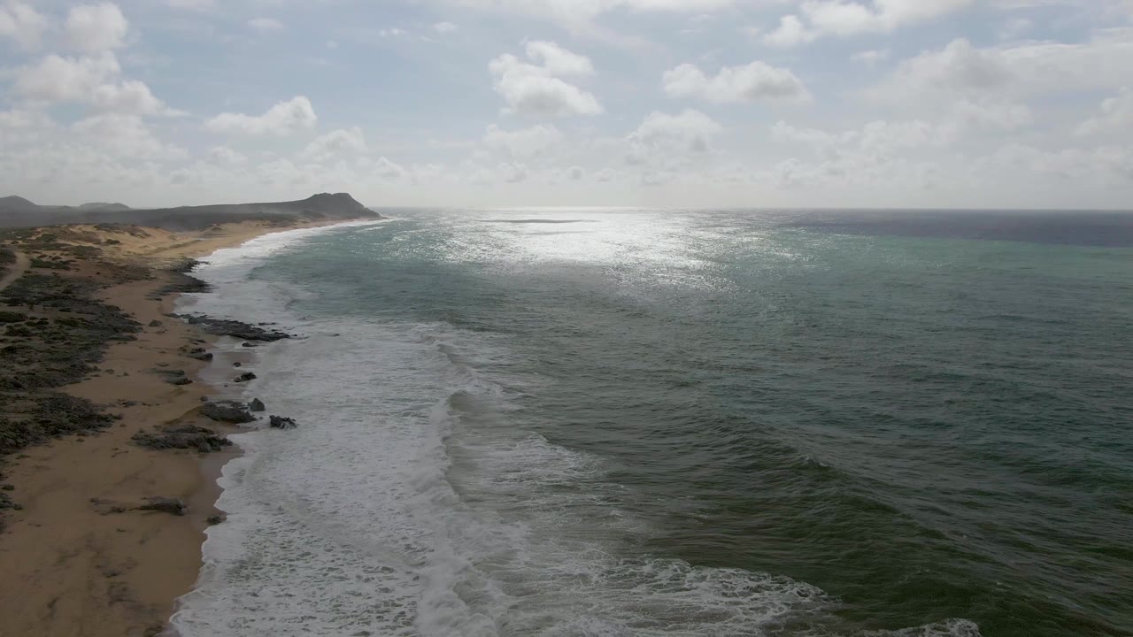 At 0:01, the Punta Gorda East Cape coast remains framed from above as a broad band of foam advances along the curved sand, with rocky patches at left, sunlit ocean at center-right, and no visible people.