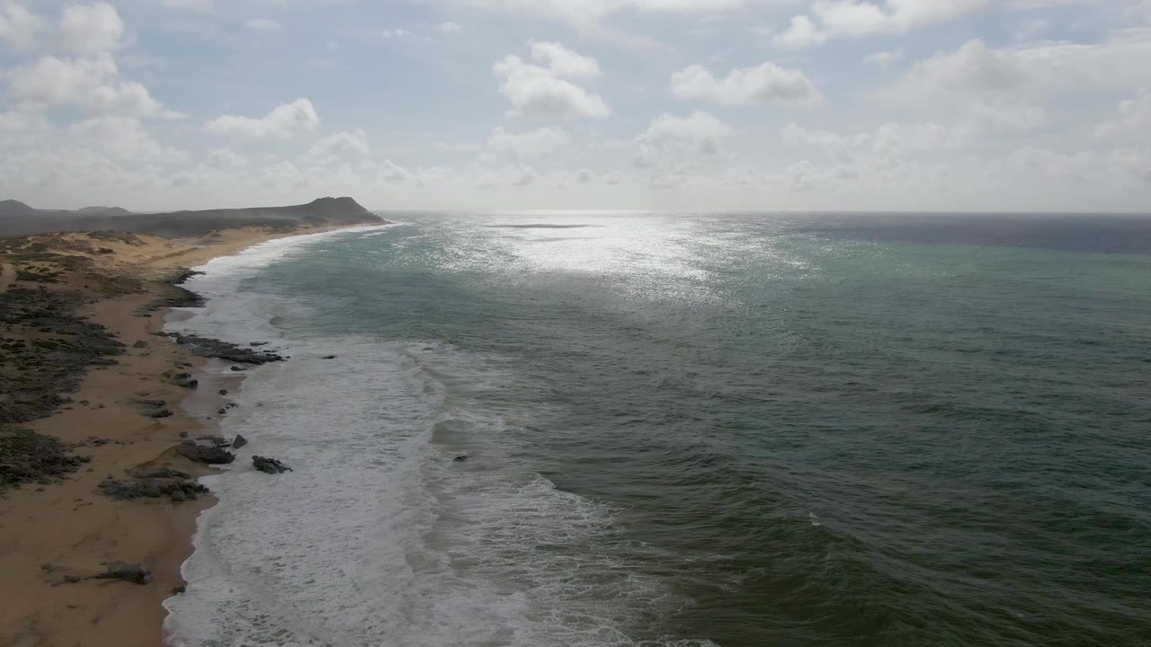 At 0:02, white surf spreads across the nearshore water beside the Punta Gorda East Cape beach, while dark rocks break up the sand, the ocean surface glints in the sun, and no visible people appear.
