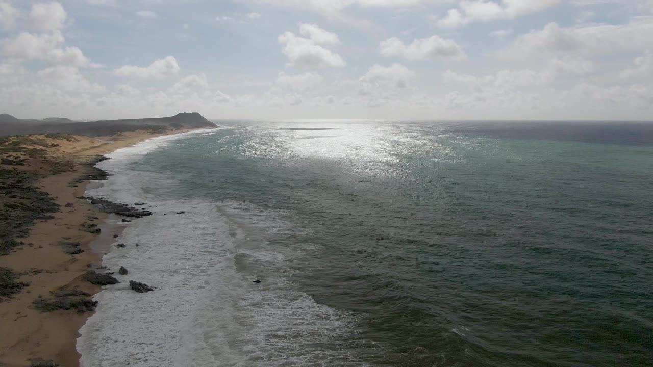 At 0:03, the aerial view shows Punta Gorda East Cape waves washing toward the sand, scattered rocks in the lower left shoreline, a bright reflective patch on the sea, and no visible people.