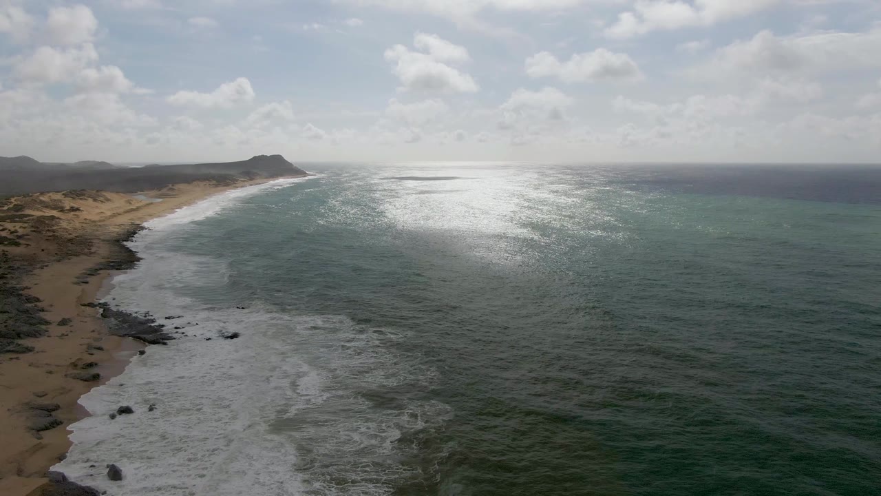 Aerial frame over Punta Gorda, East Cape, Baja California Sur, with white surf meeting a sandy beach, dark rocks near shore, bright ocean glare, distant hills, and no visible people.
