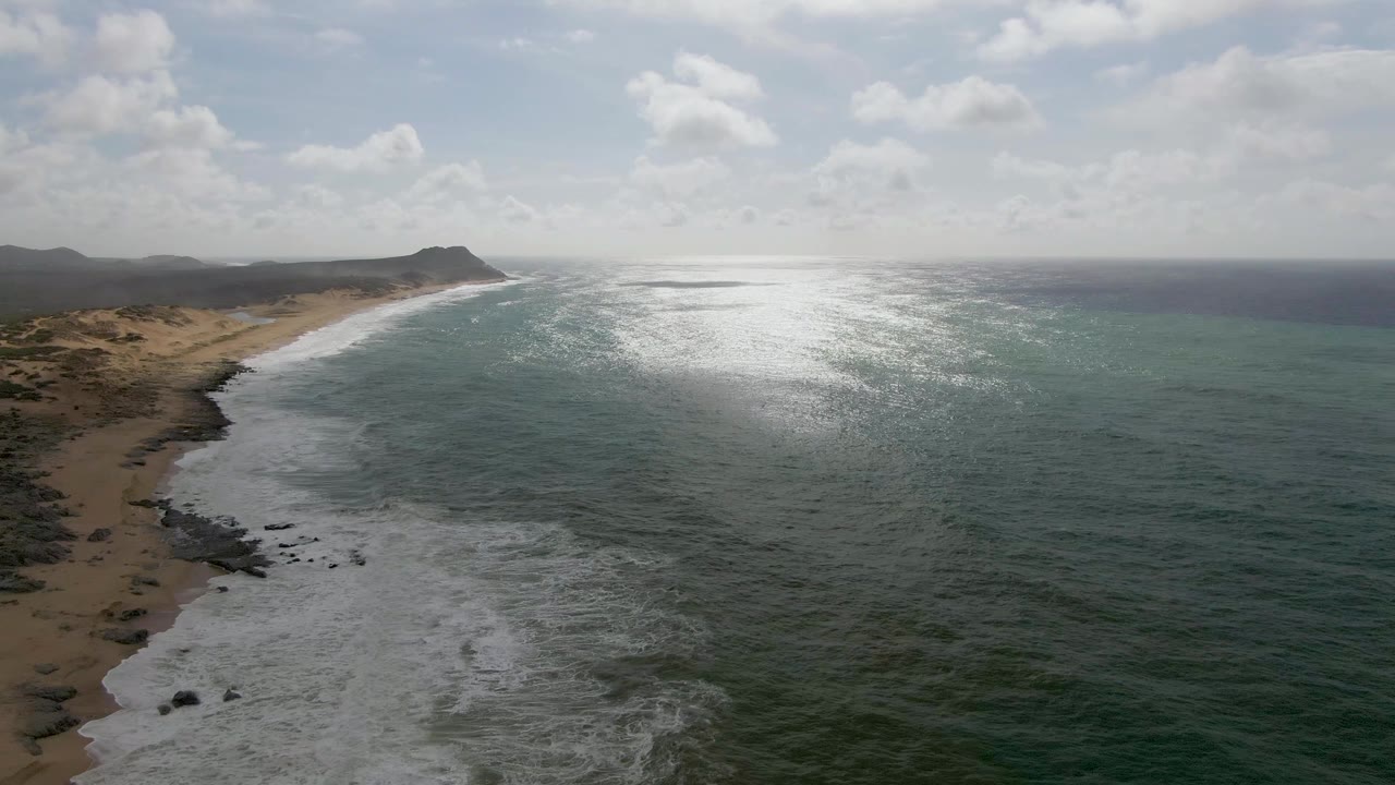 Waves break along a sandy shoreline with rocks and bright water glare.