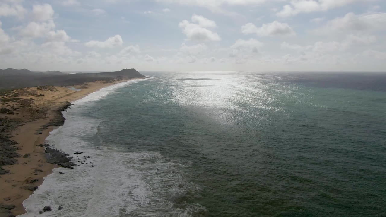 Punta Gorda aerial frame showing a long sandy beach edged by breaking white waves, scrub-covered coastal land, open green-blue ocean, a bright reflection patch, distant hills, and no visible people.