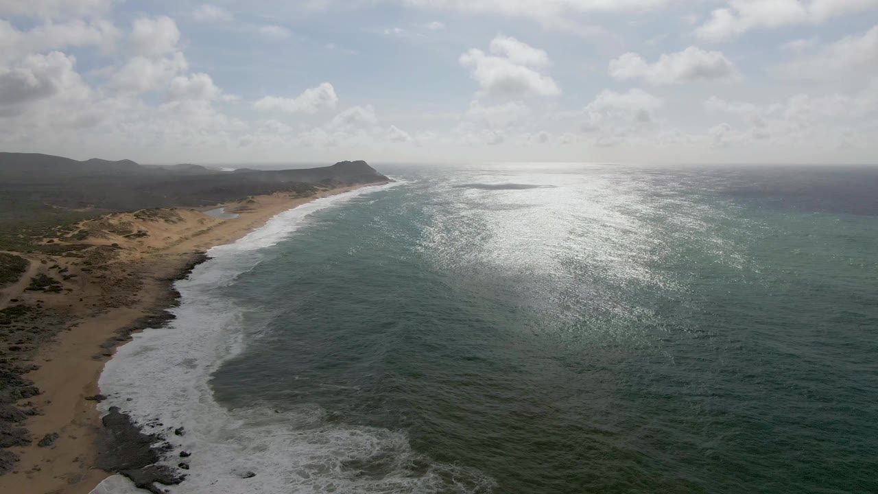 Aerial view of Punta Gorda East Cape coast with white surf along tan sand, low dunes and hills to the left, bright glare on the ocean, and no visible people.