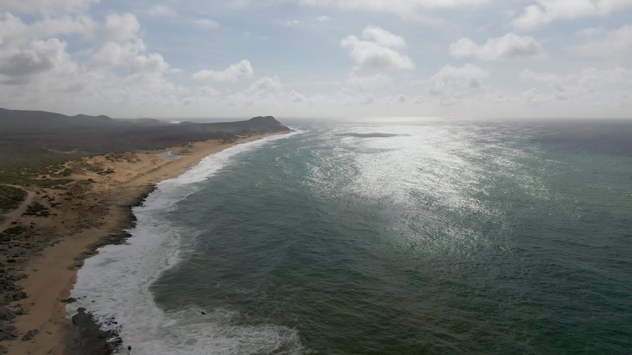 Drone view along the Punta Gorda East Cape shoreline, with breaking waves tracing the beach, dunes inland, a hazy hill line, and no visible people.