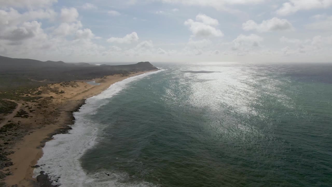 Wide aerial frame from Punta Gorda East Cape with white foam curling onto sand, dark green-blue water to the right, low brown dunes inland, and no visible people.