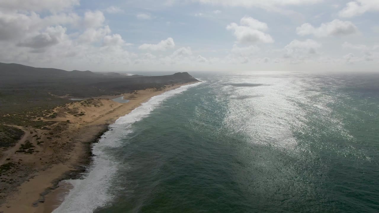 A high aerial view at Punta Gorda, Baja California Sur shows white surf curling onto a sandy beach beside green-blue water, low scrub, distant hills, scattered clouds, and no visible people.