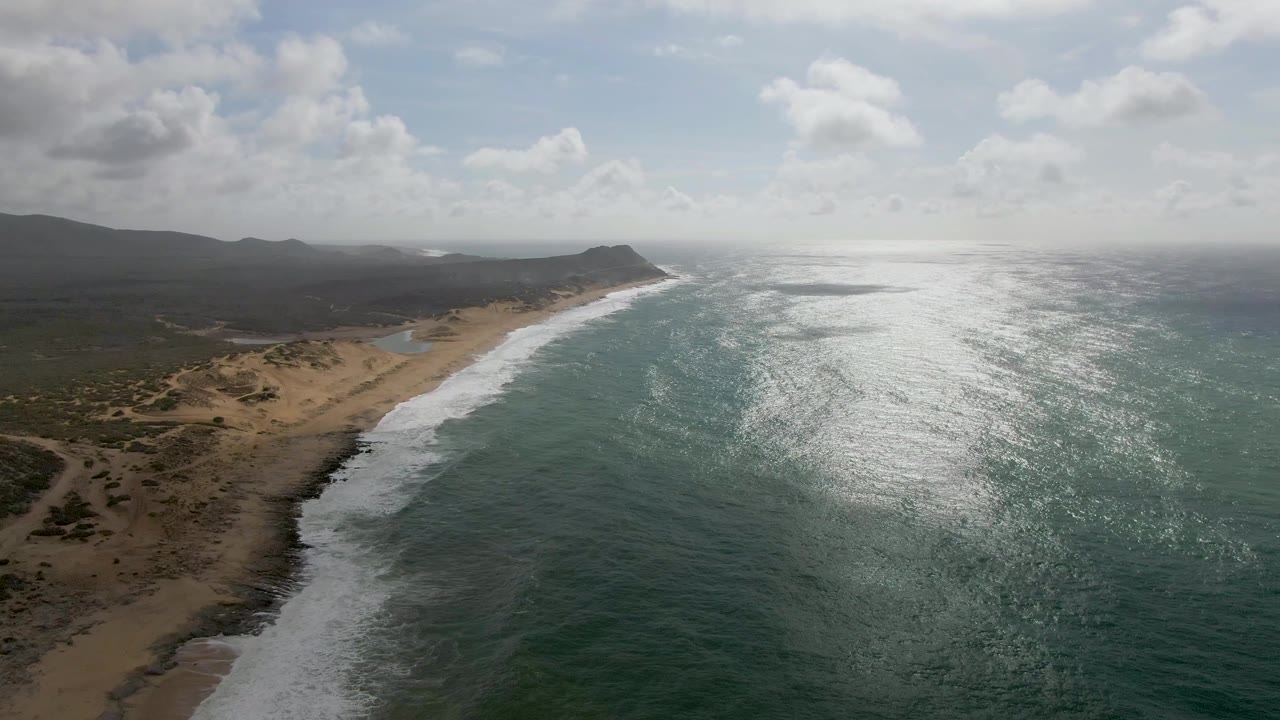 At 0:01, the Punta Gorda shoreline remains in a high oblique view as white foam advances along the sand and a broad patch of sunlight reflects across the ocean, with no visible people.