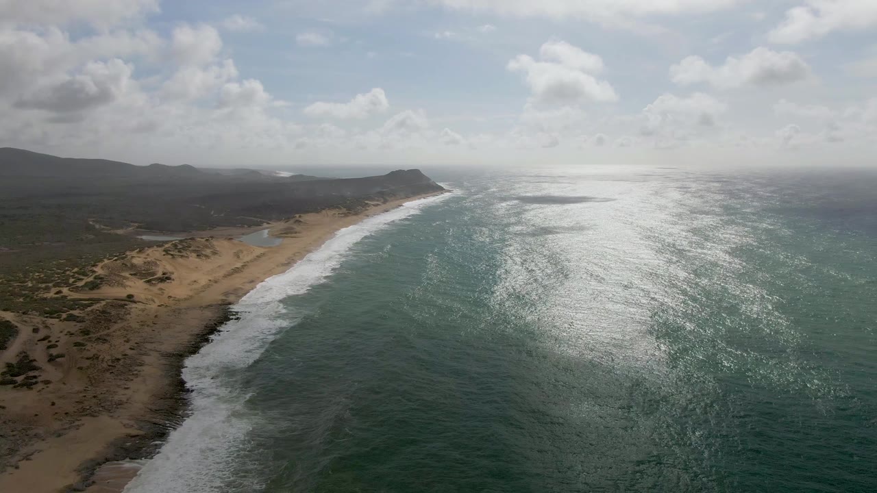 At 0:02, foamy waves spread across the near shore at Punta Gorda while the sandy beach and scrubland curve toward low hills under scattered clouds, with no visible people.
