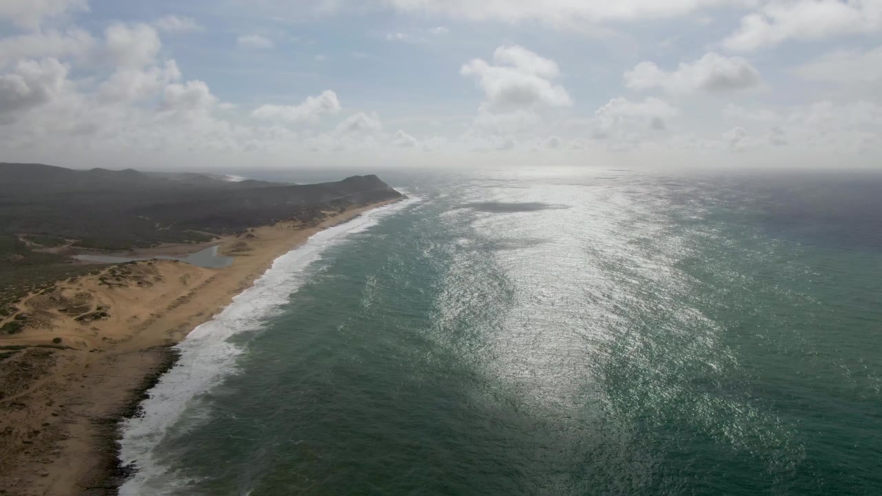At 0:00, an aerial view over Punta Gorda East Cape shows teal ocean water, white surf, sandy shore, dry hills, scattered clouds, and no visible people.